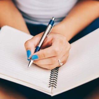 A woman wiht blue nails and a diamond ring on her middle finger writing in a spiral notebook in her lap with a steel pen.