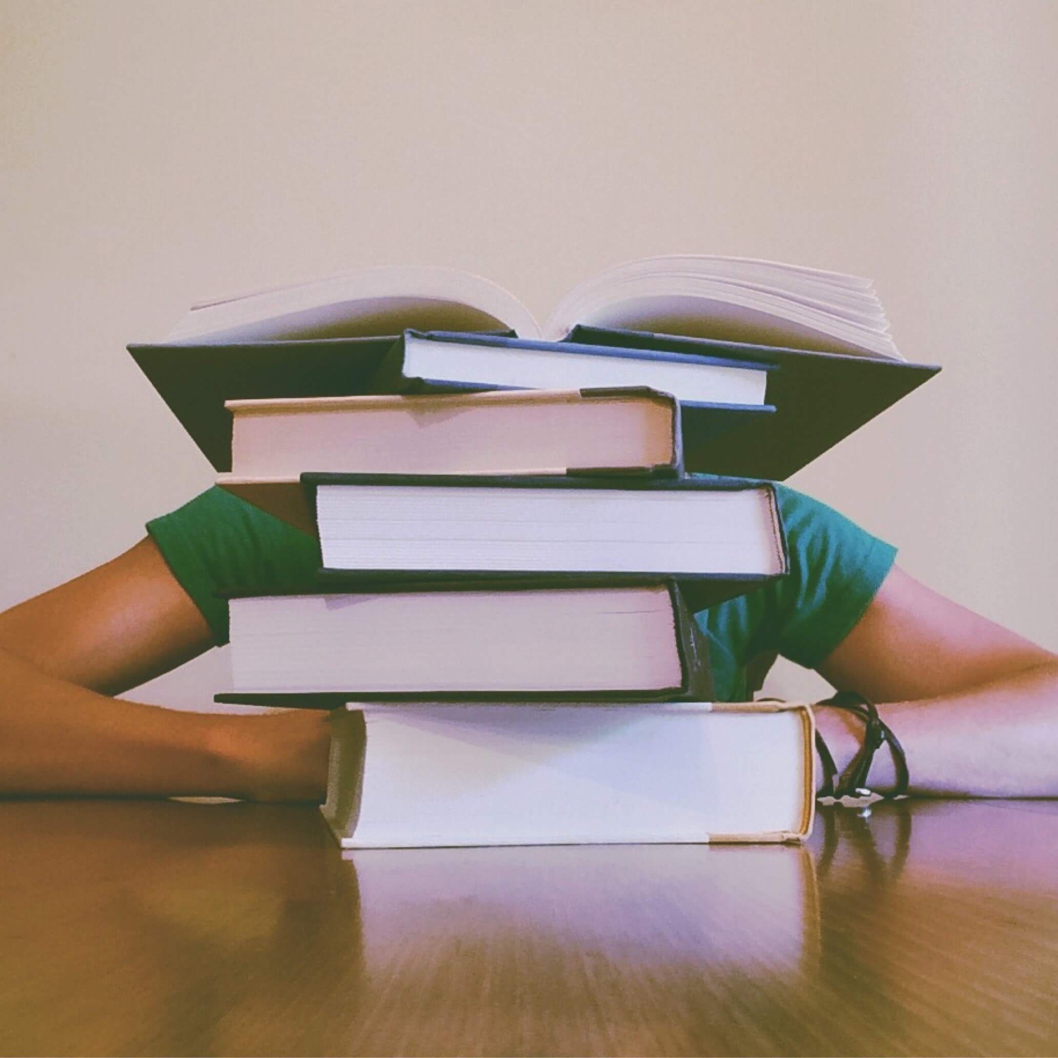 A person in a green shirt leaning on the table behind a stack of books with the book on top opened in the middle.