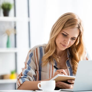 Blonde woman in a striped white-blue-golden shirt in a sunny room writing in a notebook before a laptop with a coffee cup.