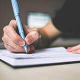 A hand of a person in a black shirt writing in a notebook with a blue pen.
